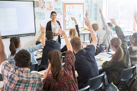 An image of a teacher at the front of a classroom and students sitting at desks with their hands up