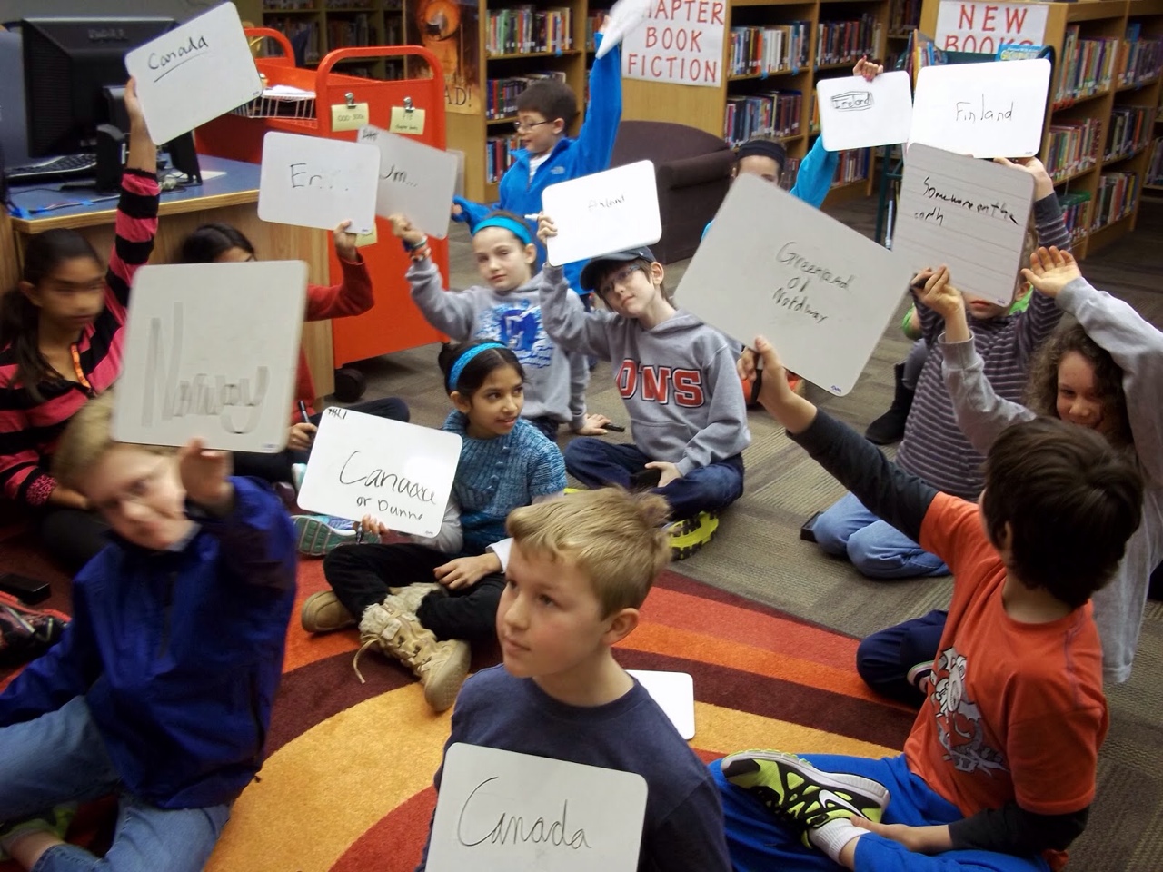 An image of a class sitting on the floor all holding up mini whiteboards with answers written on them